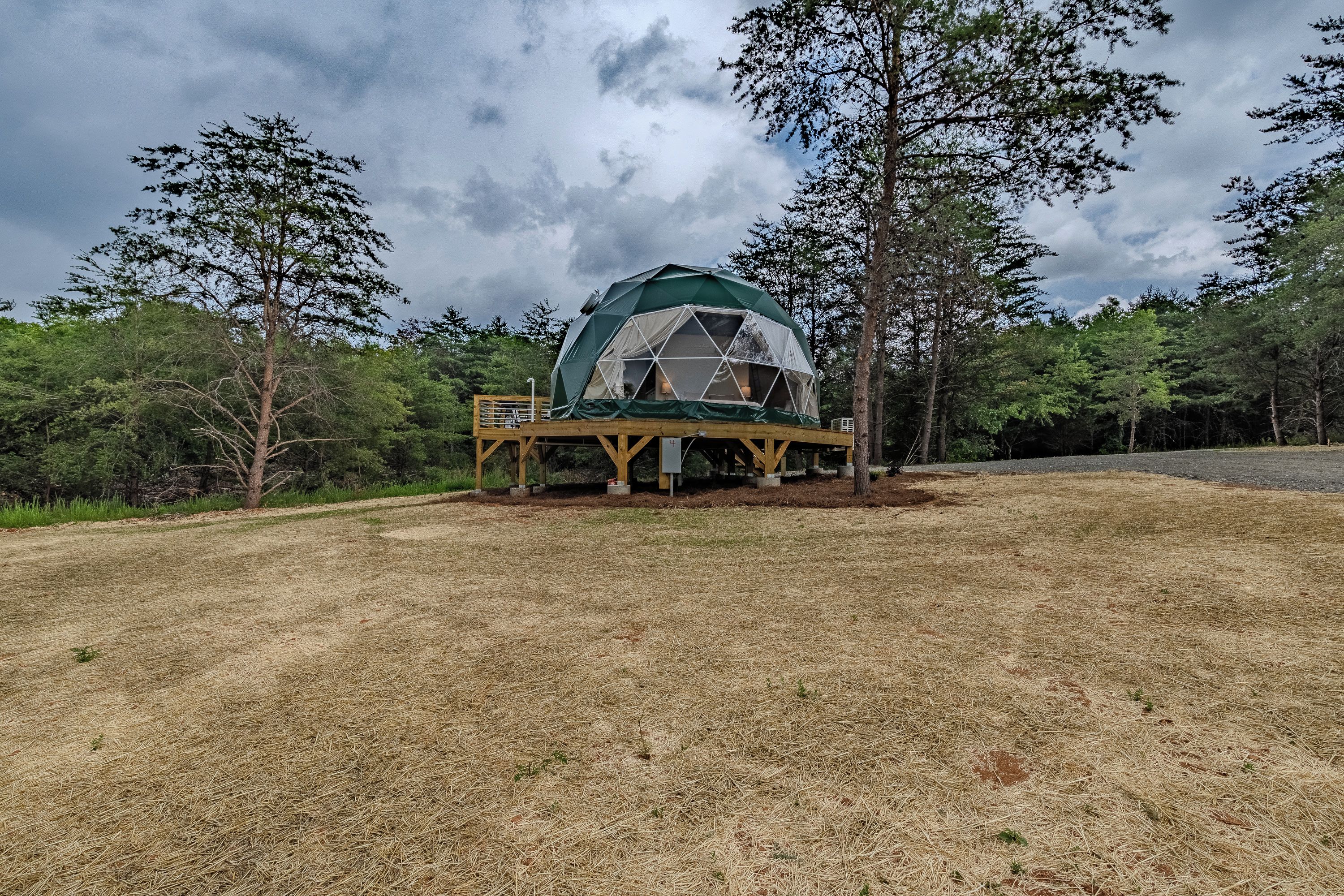 A geodesic dome structure with a wooden deck, surrounded by trees and grassy terrain.