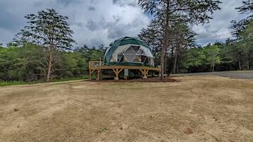 A geodesic dome structure with a wooden deck, surrounded by trees and grassy terrain.