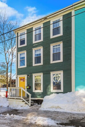 A two-story building with a green exterior and a yellow door, surrounded by snow. - Colonial Row 3B, Off-Street Parking, Private Patio (St. John's)