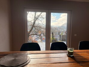 Une pièce avec une table en bois, deux chaises, un verre de lait et une vue par la fenêtre sur un paysage enneigé, avec des maisons et des arbres.