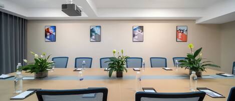 Uma sala de conferências com uma mesa comprida, cadeiras, plantas em vasos, garrafas de água e quadros nas paredes.