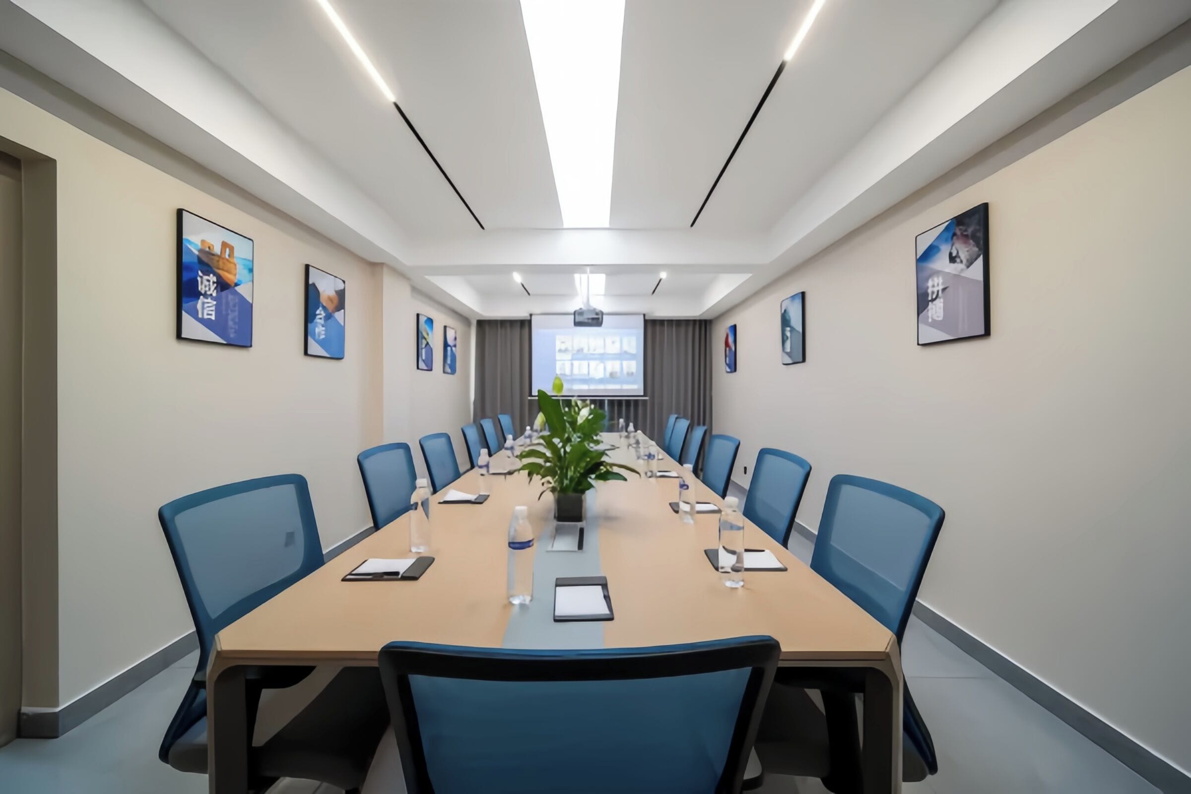 A conference room with a long table, blue chairs, a projector screen, and framed pictures on the walls.