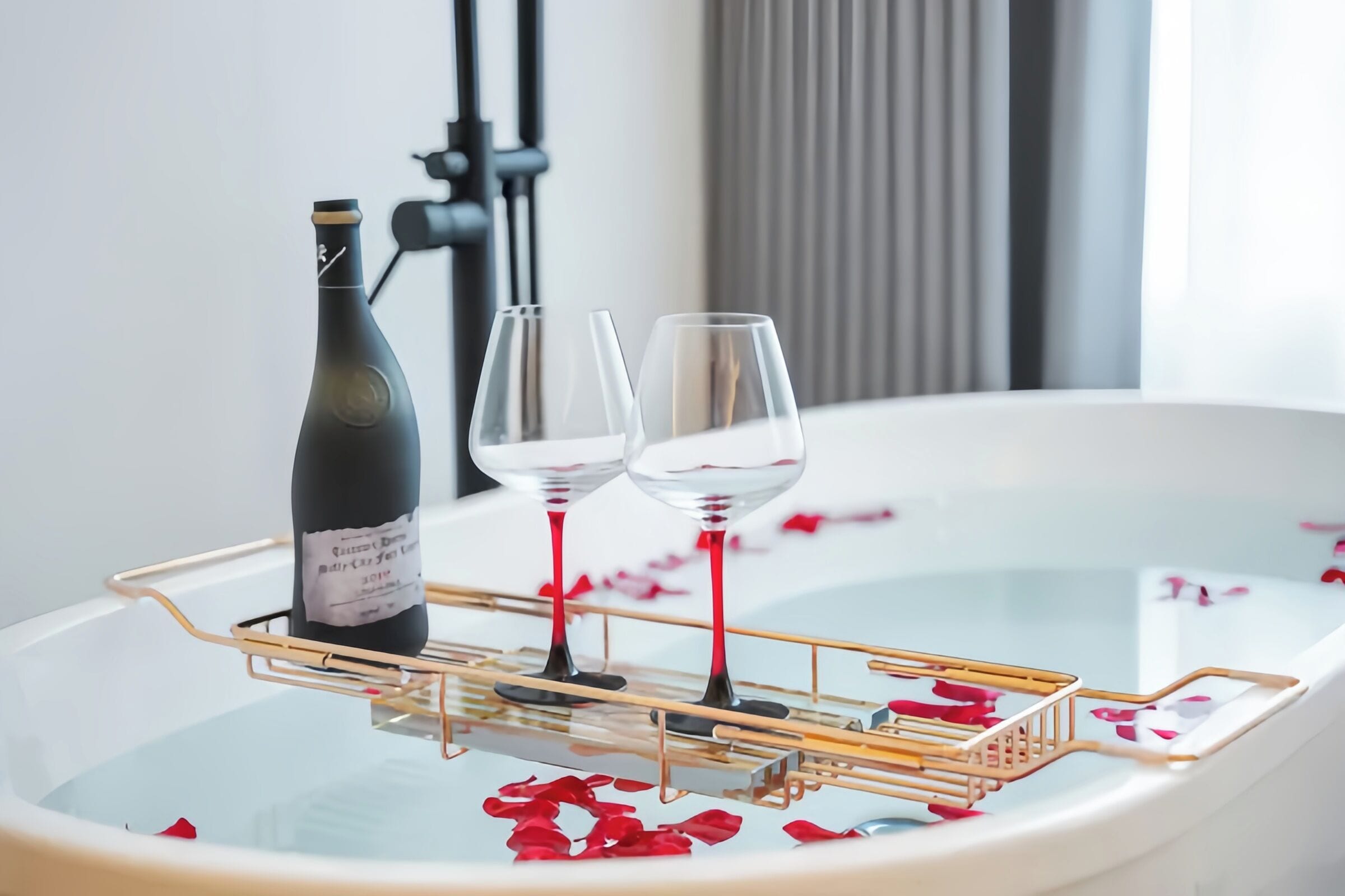 A bathtub with rose petals and two wine glasses, a bottle of wine, and a gold tray.