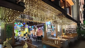 Een terras met tafels en banken, slingers met lichtjes erboven, en diverse planten en decoraties.