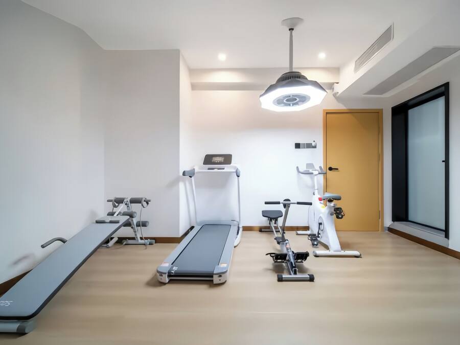 Un gimnasio moderno con cinta de correr, elíptica y bicicleta estática, suelo de madera y una lámpara en el techo.