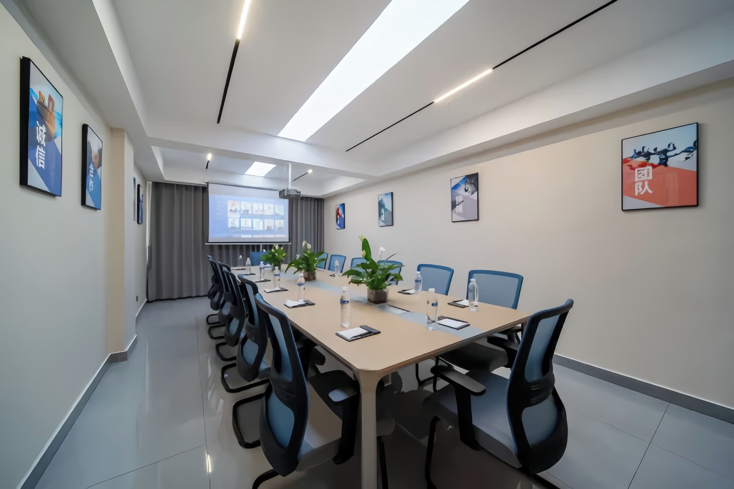 A conference room with a long table, chairs, a projector screen, and framed pictures on the walls.