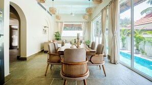 A dining area with a round table, wicker chairs, and a view of a swimming pool. - Staymaster White Castle-4BHK (Vagator)