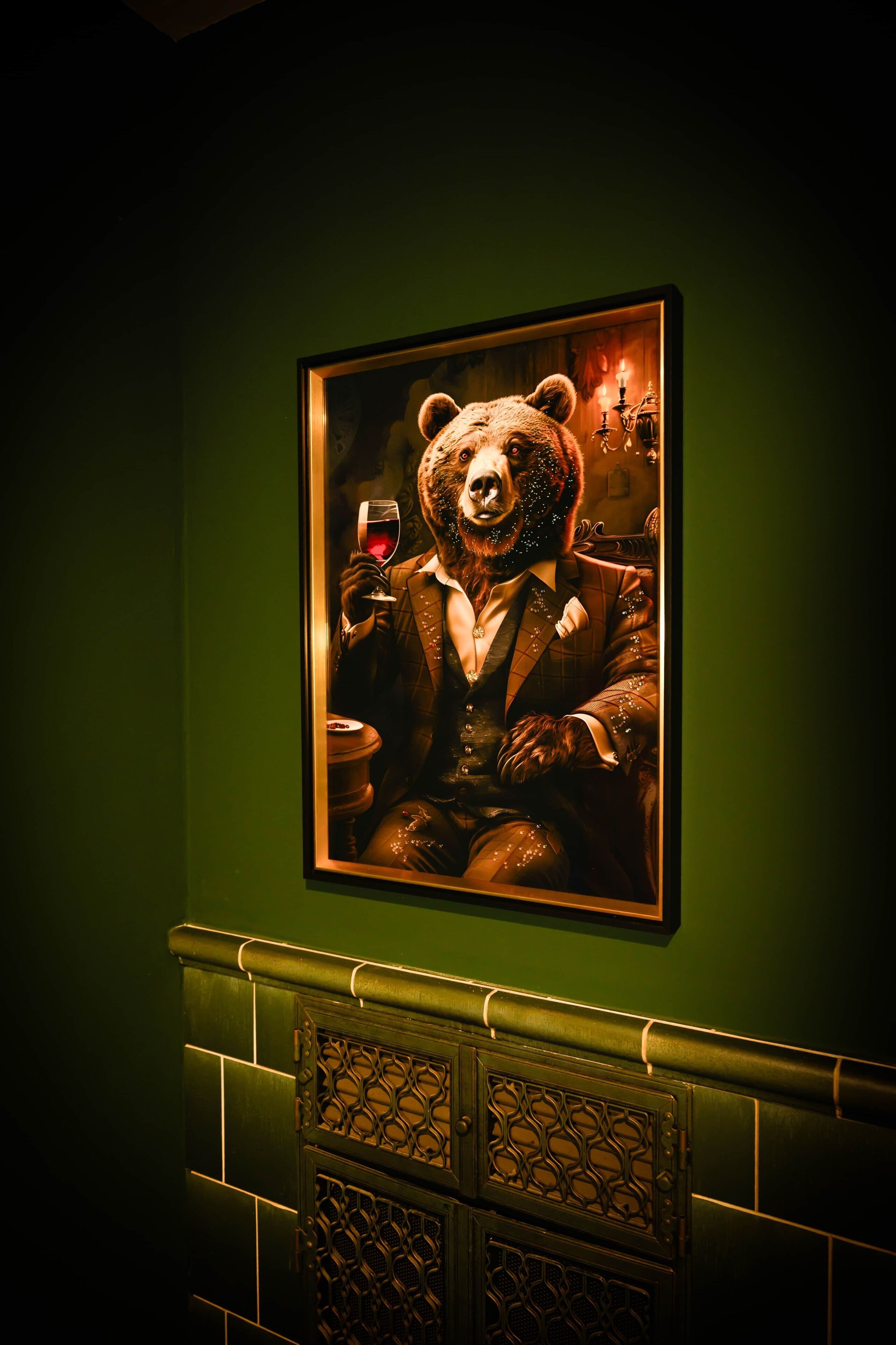 A framed portrait of a bear in a suit holding a glass of wine, set against a green wall with a decorative fireplace below.
