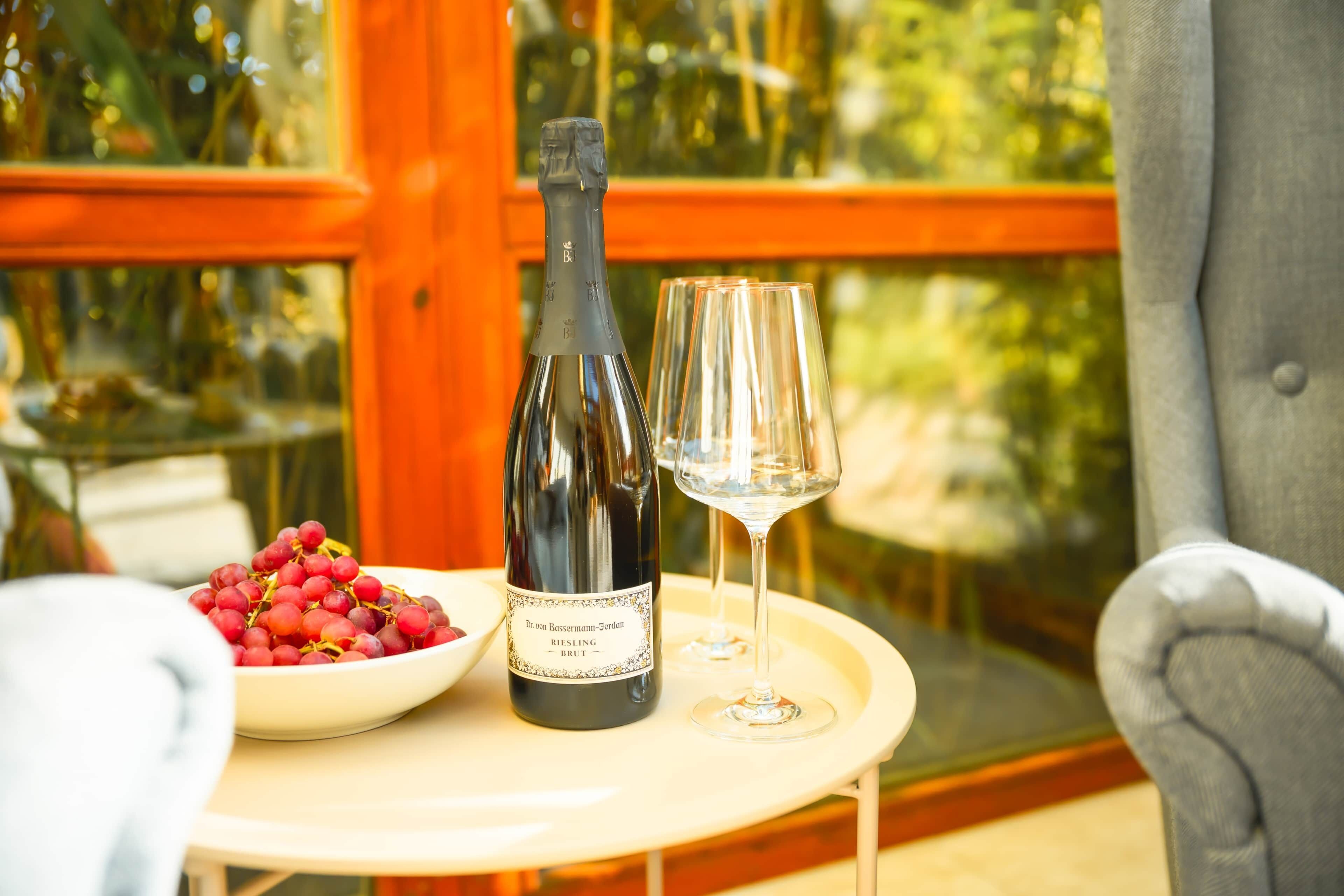 A bottle of wine, a glass, and a bowl of grapes on a table by a window.
