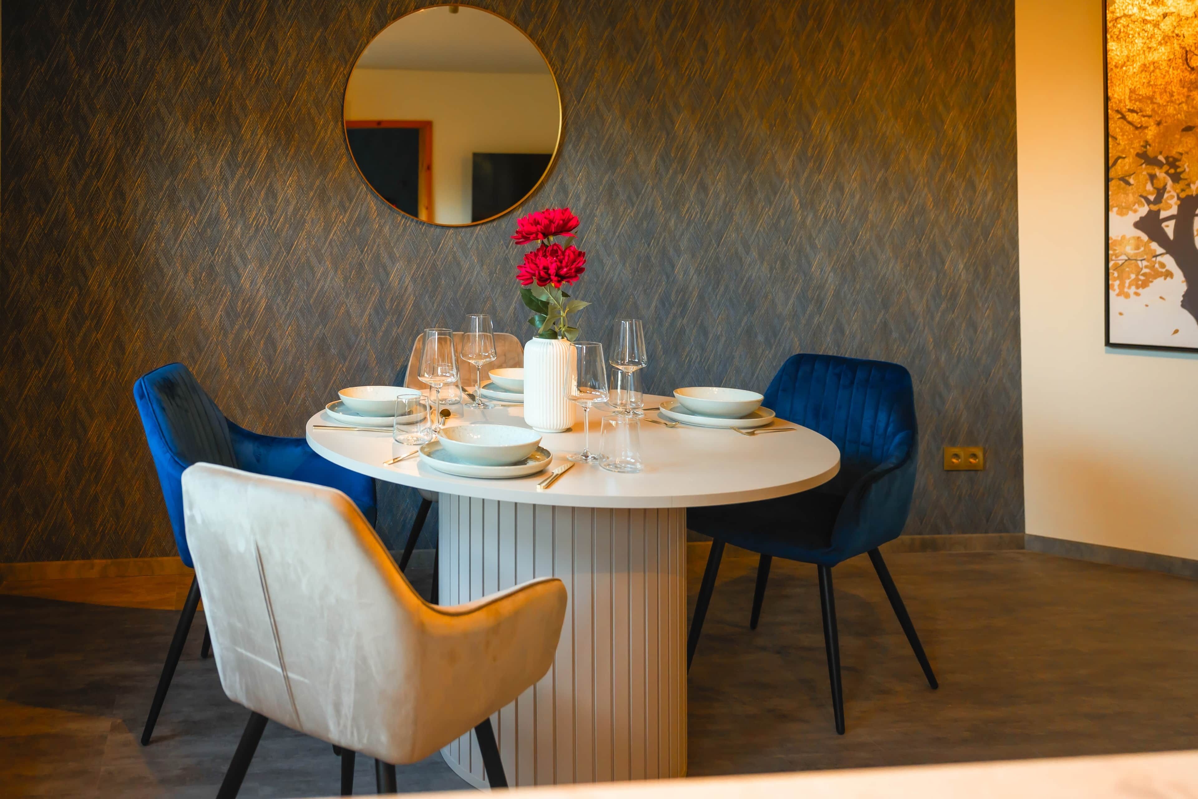 Ein Essbereich mit einem runden Tisch, zwei blauen Stühlen und einer weißen Vase mit roten Blumen.