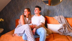 A couple sitting on an orange sofa in a room with a herringbone-patterned wall and a round mirror above the sofa. - VIERER - Cozy Apartment XL (Spalt)