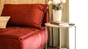 A red velvet sofa with a round mirror above it, a small white vase with flowers on a side table, and a beige curtain in the background. - DREIER - Cozy Apartment (Spalt)