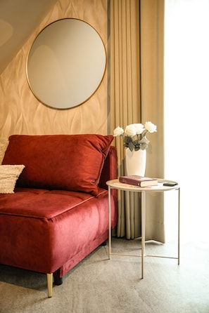 A red velvet sofa with a round mirror above it, a small white vase with flowers on a side table, and a beige curtain in the background.