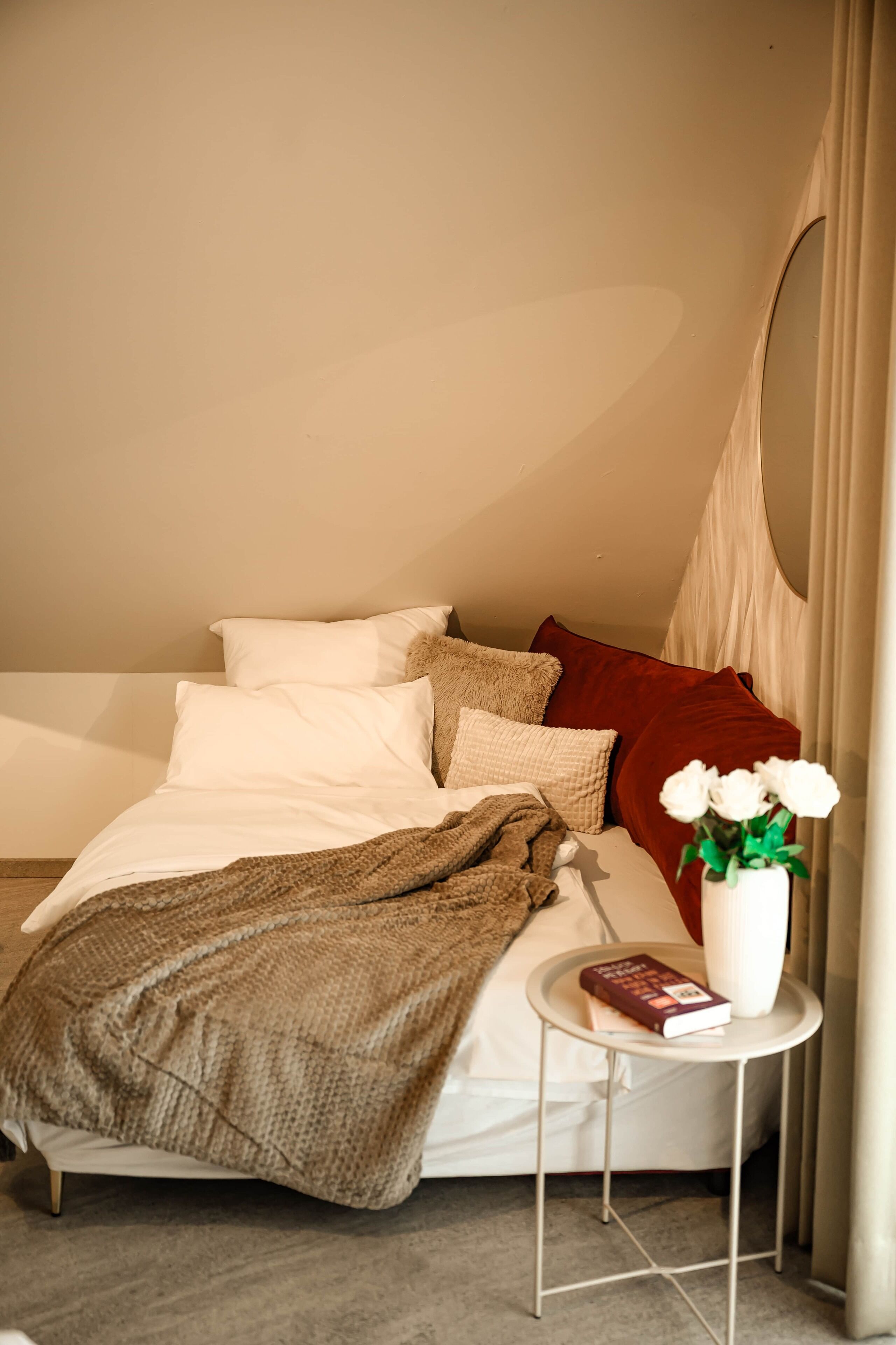 A neatly made bed with white and brown bedding, a red pillow, and a small bedside table with a book and a vase of flowers.