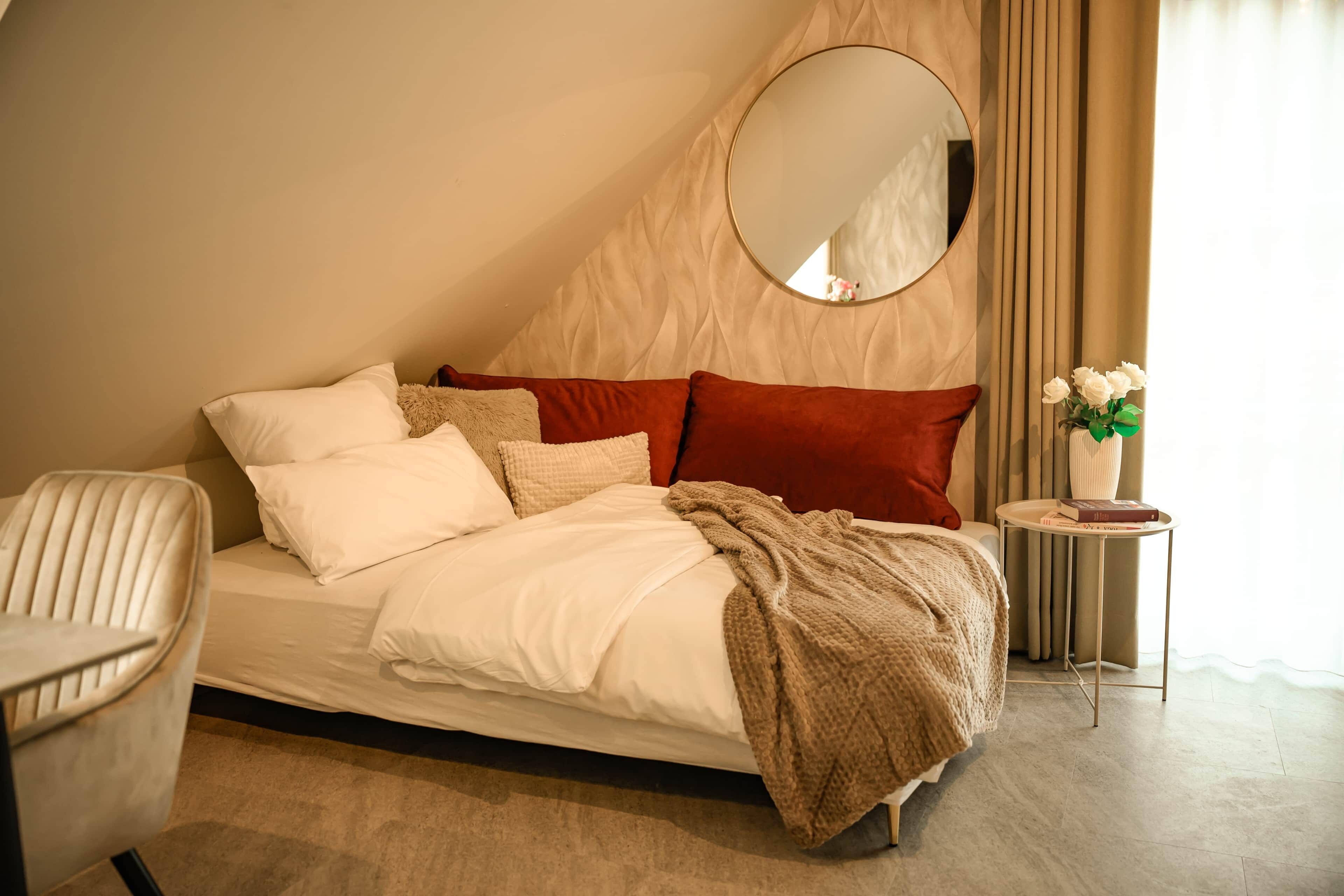 Un dormitorio con cama, una silla, una mesita de noche con un florero y un espejo redondo en la pared.