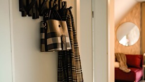 A hallway with a coat rack holding a plaid jacket and a checkered bag. A red couch and a round mirror are visible in the background. - DREIER - Cozy Apartment (Spalt)