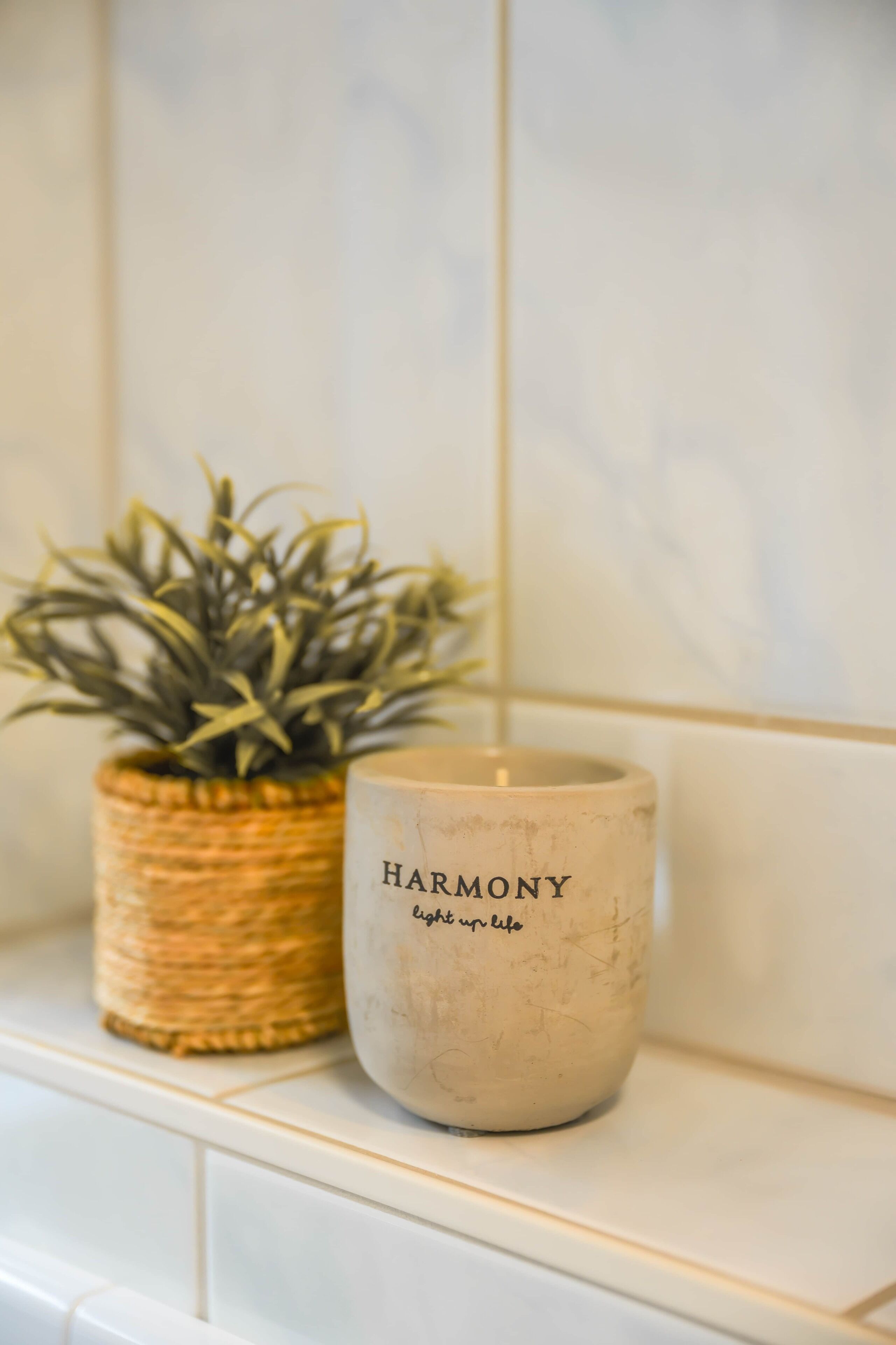 A candle labeled "HARMONY" and a potted plant on a white tiled surface.