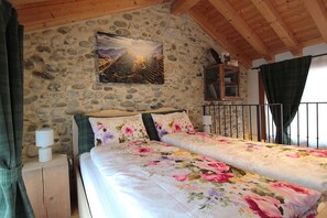 A bedroom with a stone wall, a bed with floral bedding, a nightstand with a lamp, and a painting above the bed.