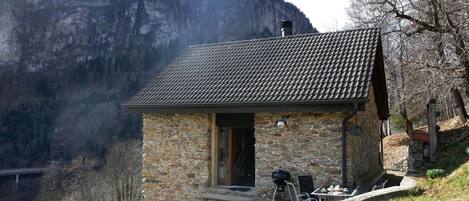 Una casa de piedra con techo de tejas, una parrilla y mobiliario exterior en una ladera con montañas al fondo.