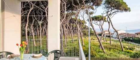 A balcony with a dining table set for two, overlooking a garden with tall trees and a view of a residential area.