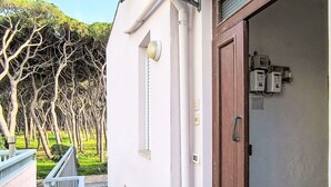 Un edificio con puerta de madera, un pequeño espacio exterior con barandilla metálica y vistas a árboles al fondo.