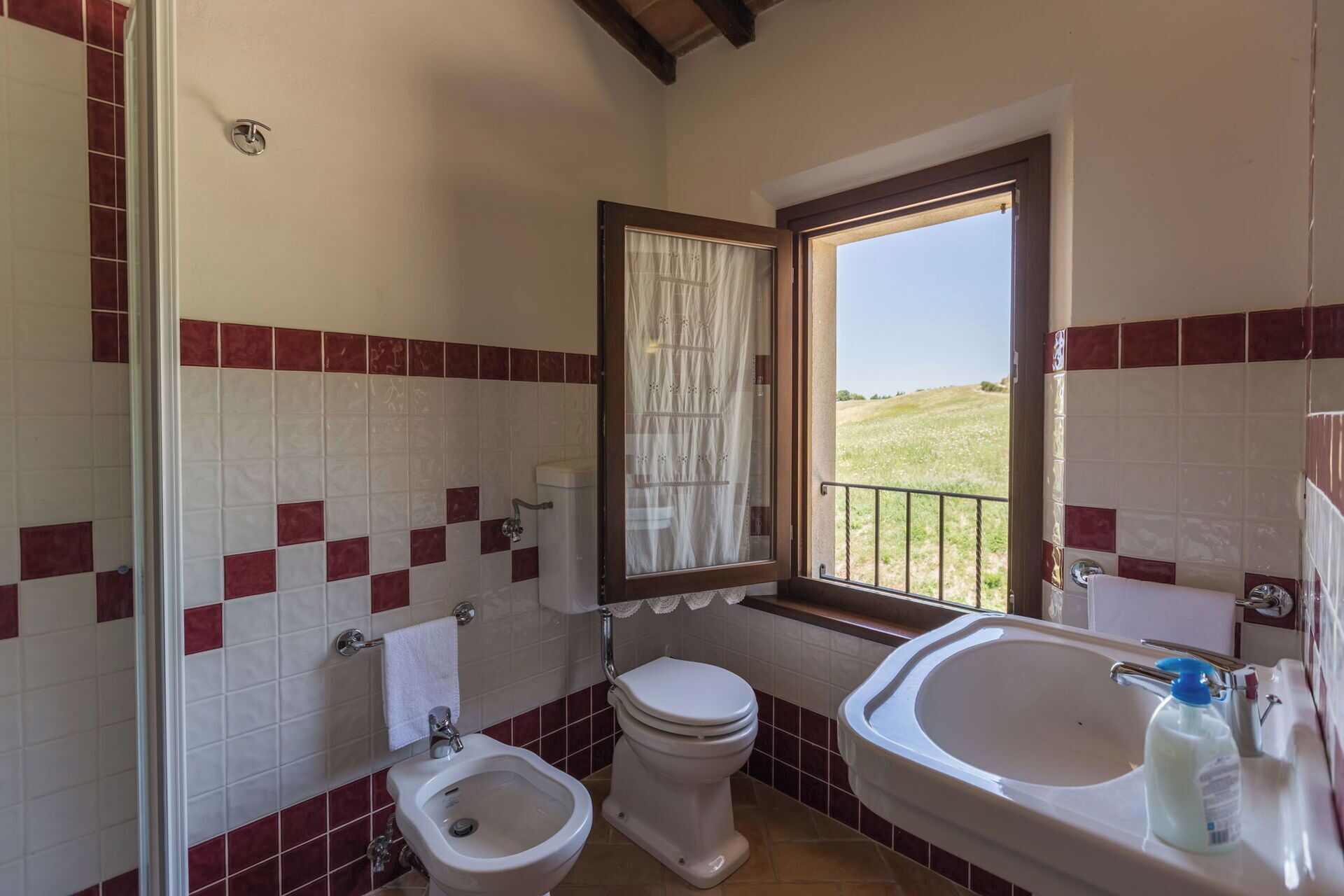 Un baño con bidé, inodoro y lavabo, con azulejos rojo y blanco y una ventana que da a un campo cubierto de hierba.