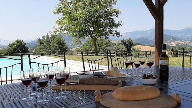 Una mesa lista para una comida, con copas de vino, una botella, pan y un bol de aceitunas, con vistas a una piscina y montañas.