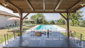 Un espacio exterior techado con piscina, sillas de descanso, sombrillas y una mesa lista para comer.