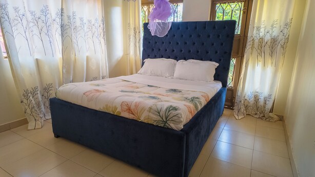 Una cama con cabecera oscura y ropa de cama floral, cortinas con motivos de árboles y suelo de baldosas.