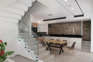 Eine moderne Küche mit einer Steinrückwand, ein Essbereich mit Holztisch und -stühlen sowie eine Glastreppe zu einer weiteren Ebene.
