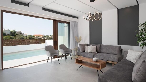 A modern living room with a grey sofa, two armchairs, a wooden coffee table, and a large sliding glass door leading to a balcony with a pool view. - O-Cyanea Villa: Luxurious 4-Bedroom Retreat with Heated Pool in Kournas (Paralia Kourna)