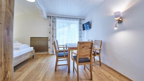 Een kamer met een houten vloer, een bed met witte lakens, een houten eettafel met stoelen, een aan de muur bevestigde televisie en een raam met gordijnen.