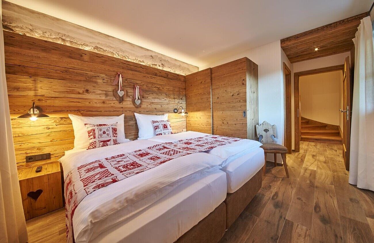 Een tweepersoonsbed met witte beddengoed en rode patronen, een houten hoofdbord, nachtlampjes en een houten vloer.