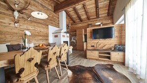 Ein rustikaler Innenraum mit Holzwänden, einem Holztisch mit Stühlen, einem Lederteppich, einem Fernseher auf einem Holzständer und einem Kamin.