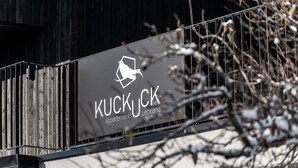 Segnaletica per 'KUCKUCK Apartments' con logo raffigurante un uccello, fissata a una ringhiera metallica su uno sfondo sfocato di rami spogli.