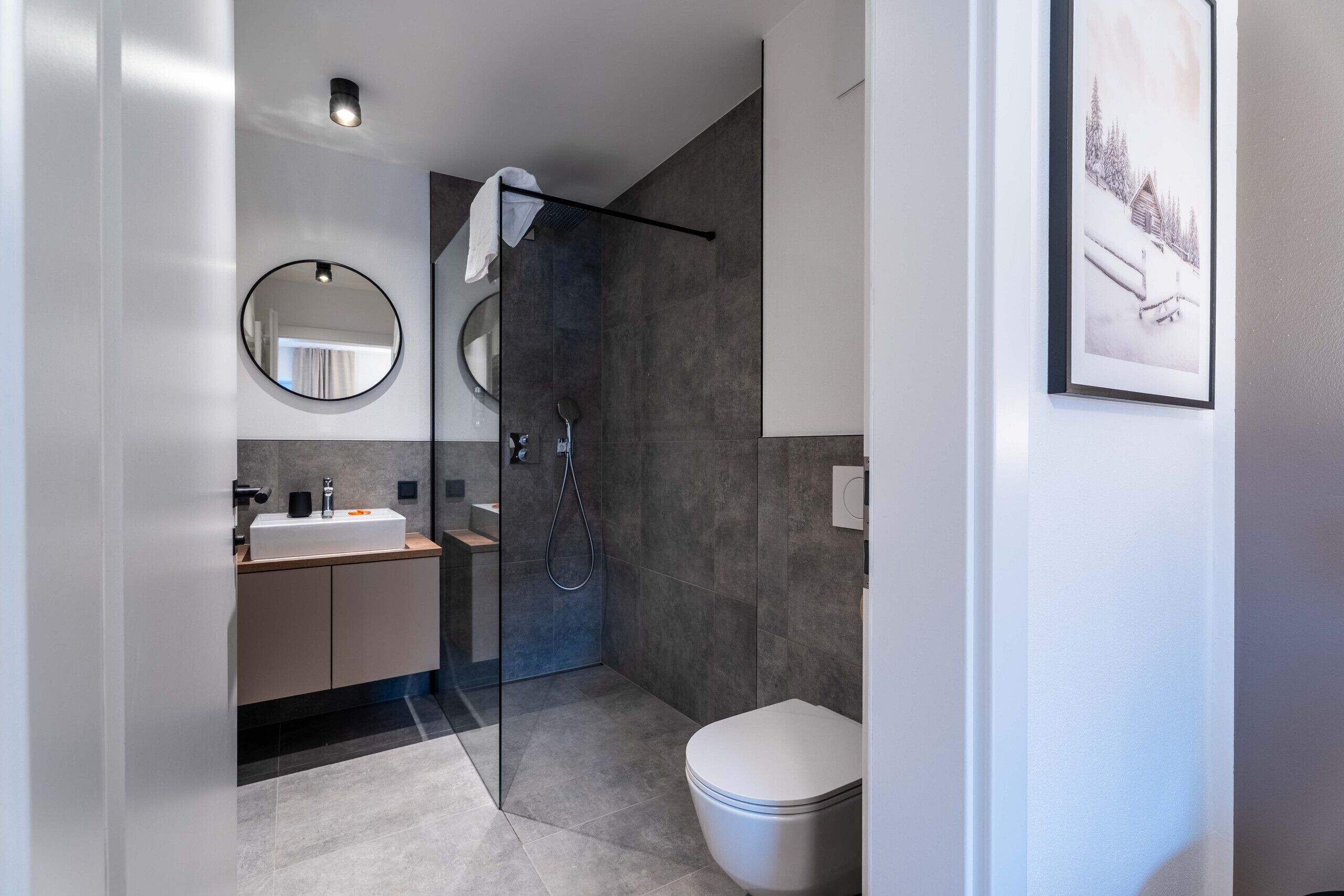 A modern bathroom with a glass-enclosed shower, a round mirror, a white toilet, and a framed picture on the wall.