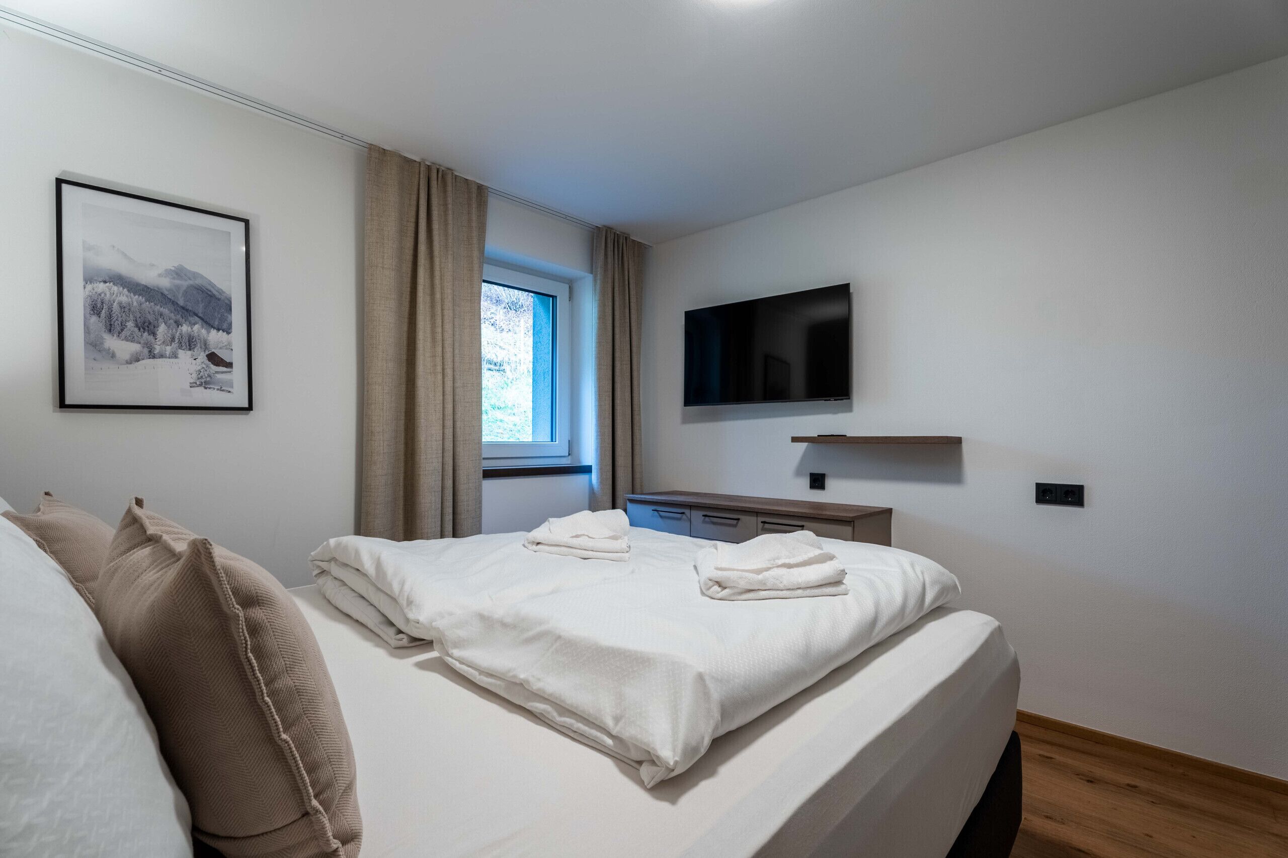 Una camera d'albergo con un letto, una TV fissata al muro, una finestra con tende e un quadro appeso alla parete.