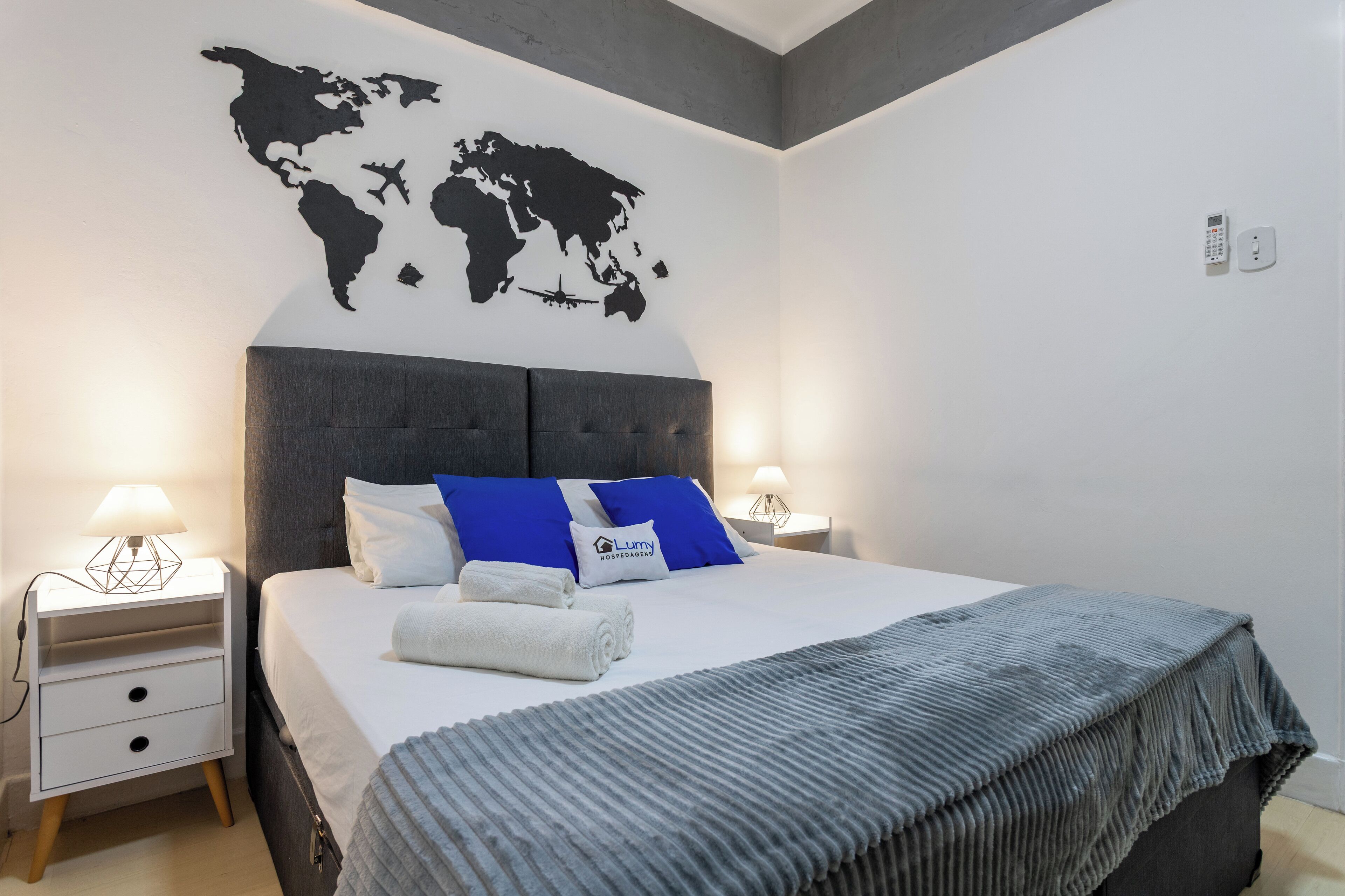 A neatly made bed with a grey blanket, a world map on the wall, and a small bedside table with a lamp.