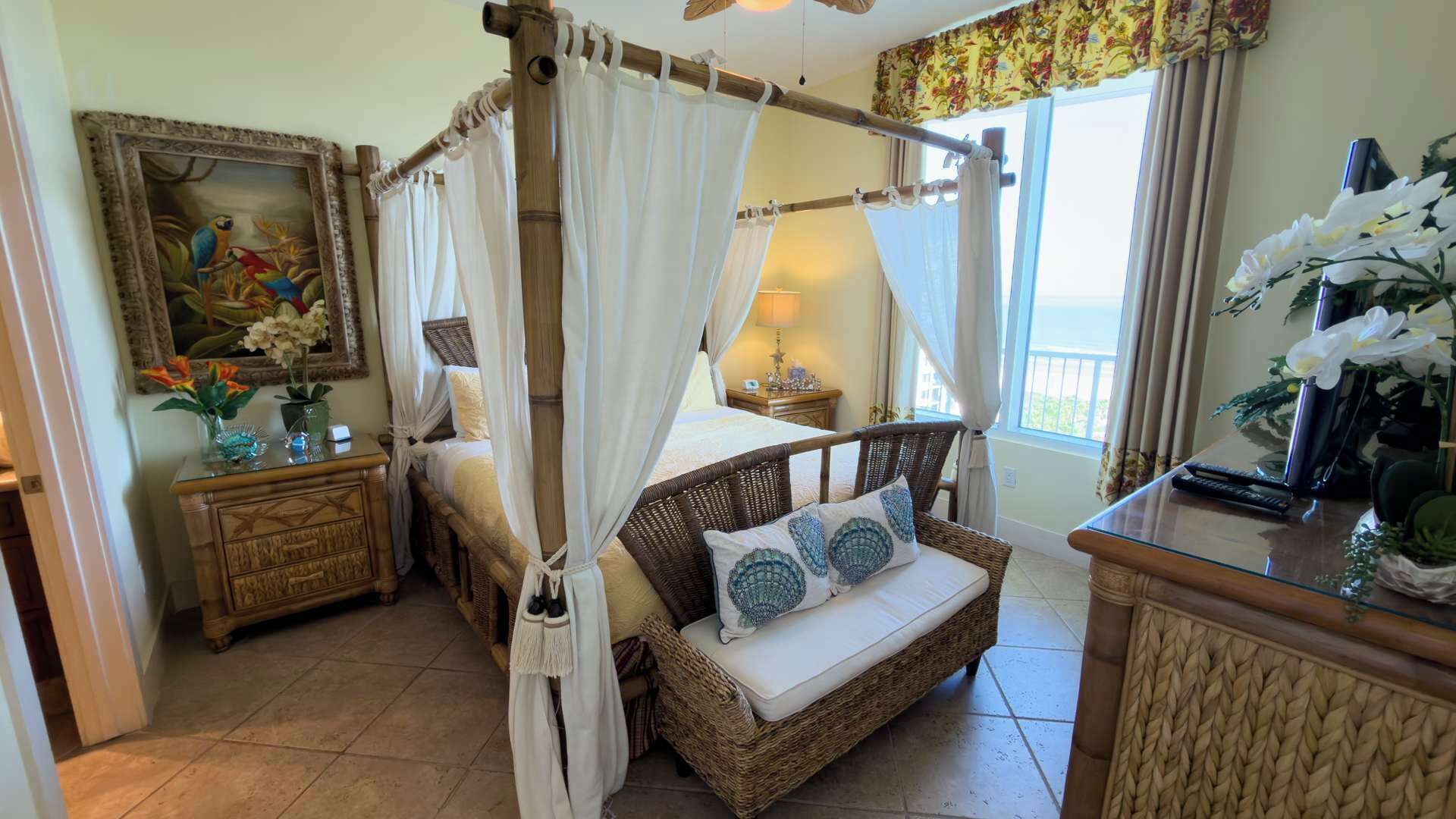 A room with a four-poster bed, a wicker sofa, a nightstand, a painting, and a view of the outdoors.