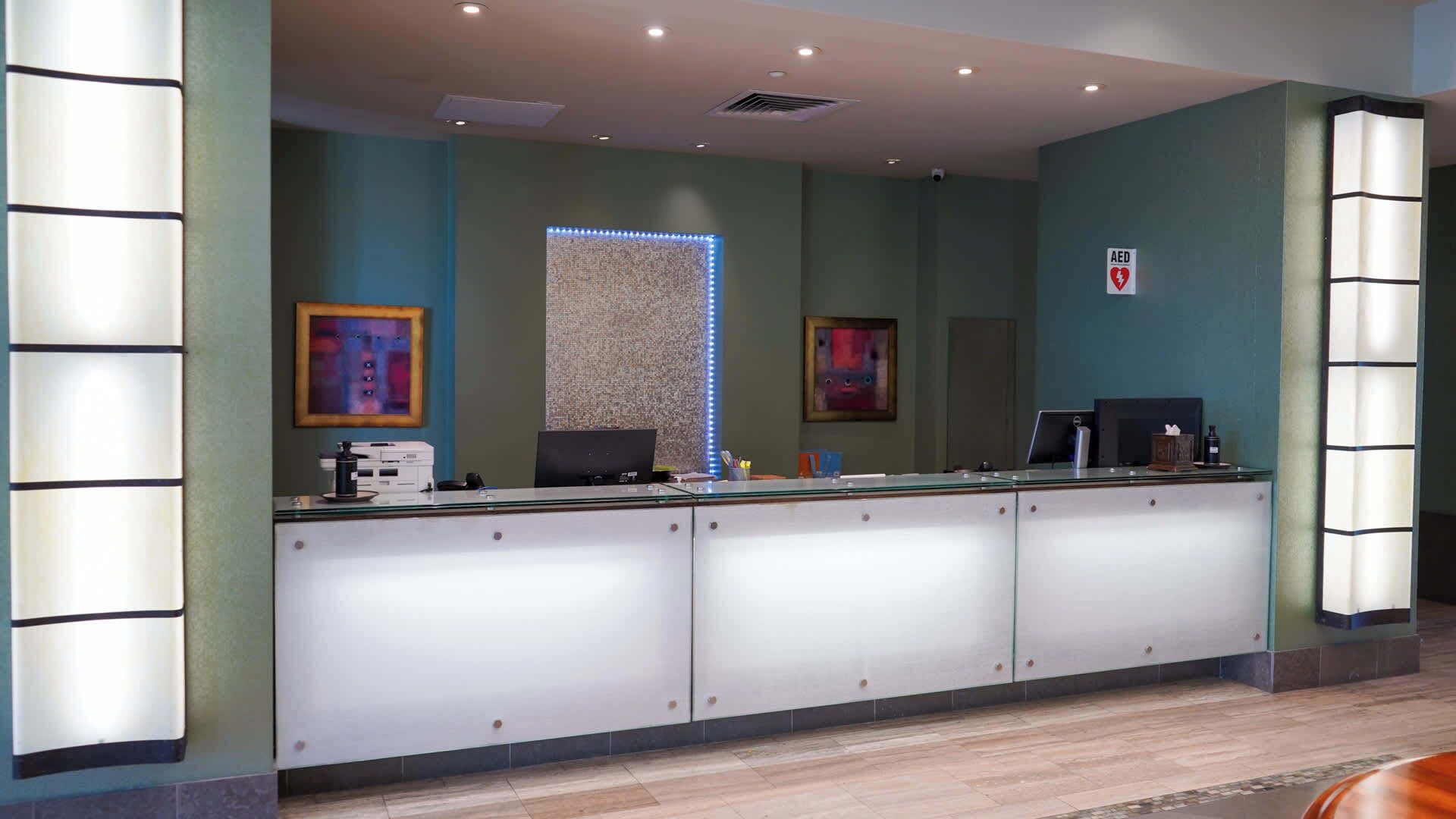 A reception area with a glass counter, two computer monitors, and a wall-mounted AED sign.
