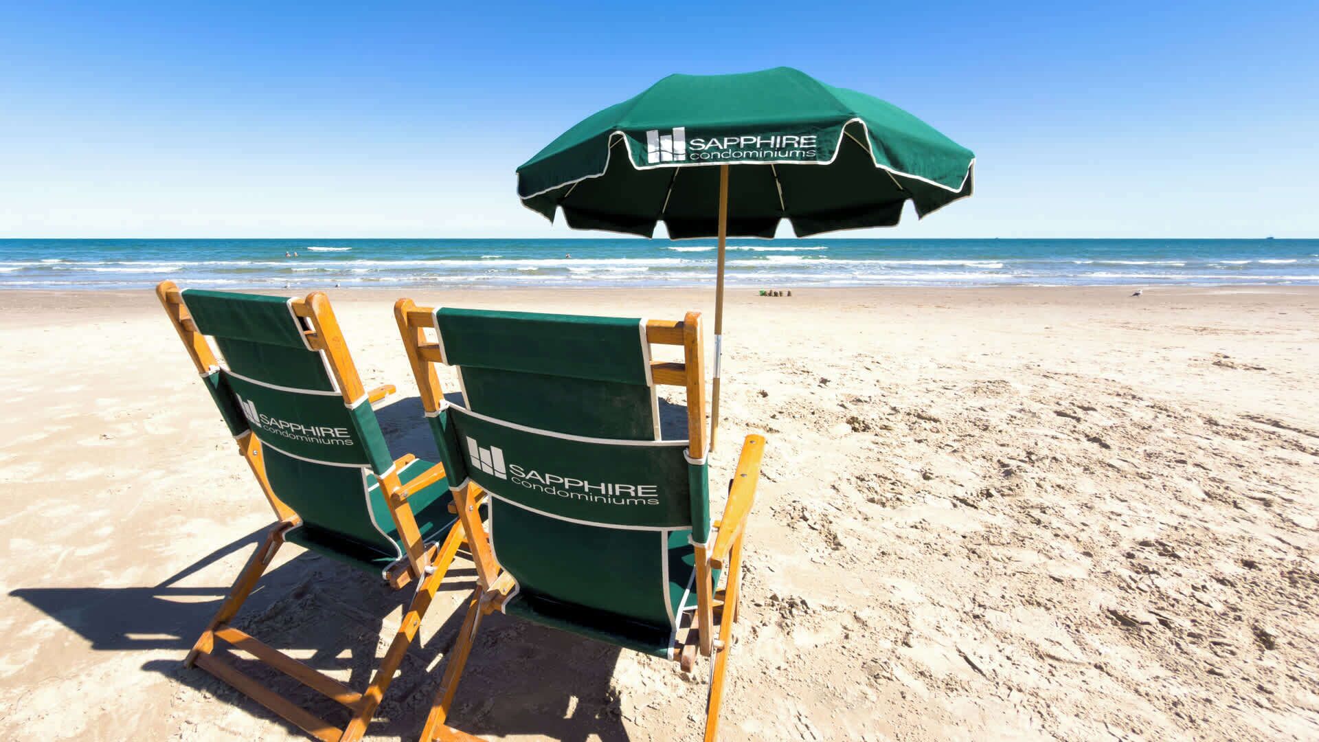 Two green lounge chairs with wooden frames and a matching green umbrella on a sandy beach.