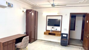 Quarto com escrivaninha de madeira, uma cadeira, guarda-roupa, televisão na parede e ventilador de teto.
