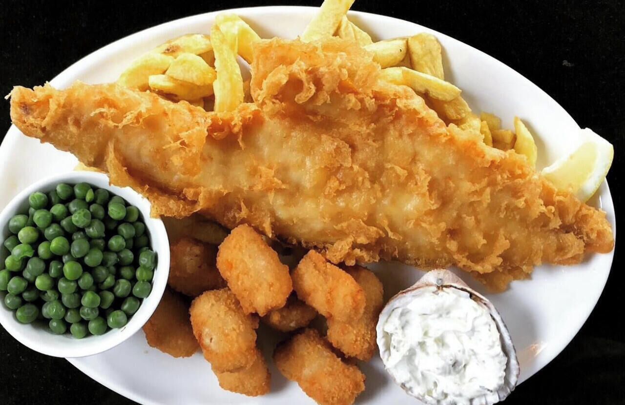 A plate with a large fish and chips, tater tots, peas, and a side of tartar sauce.