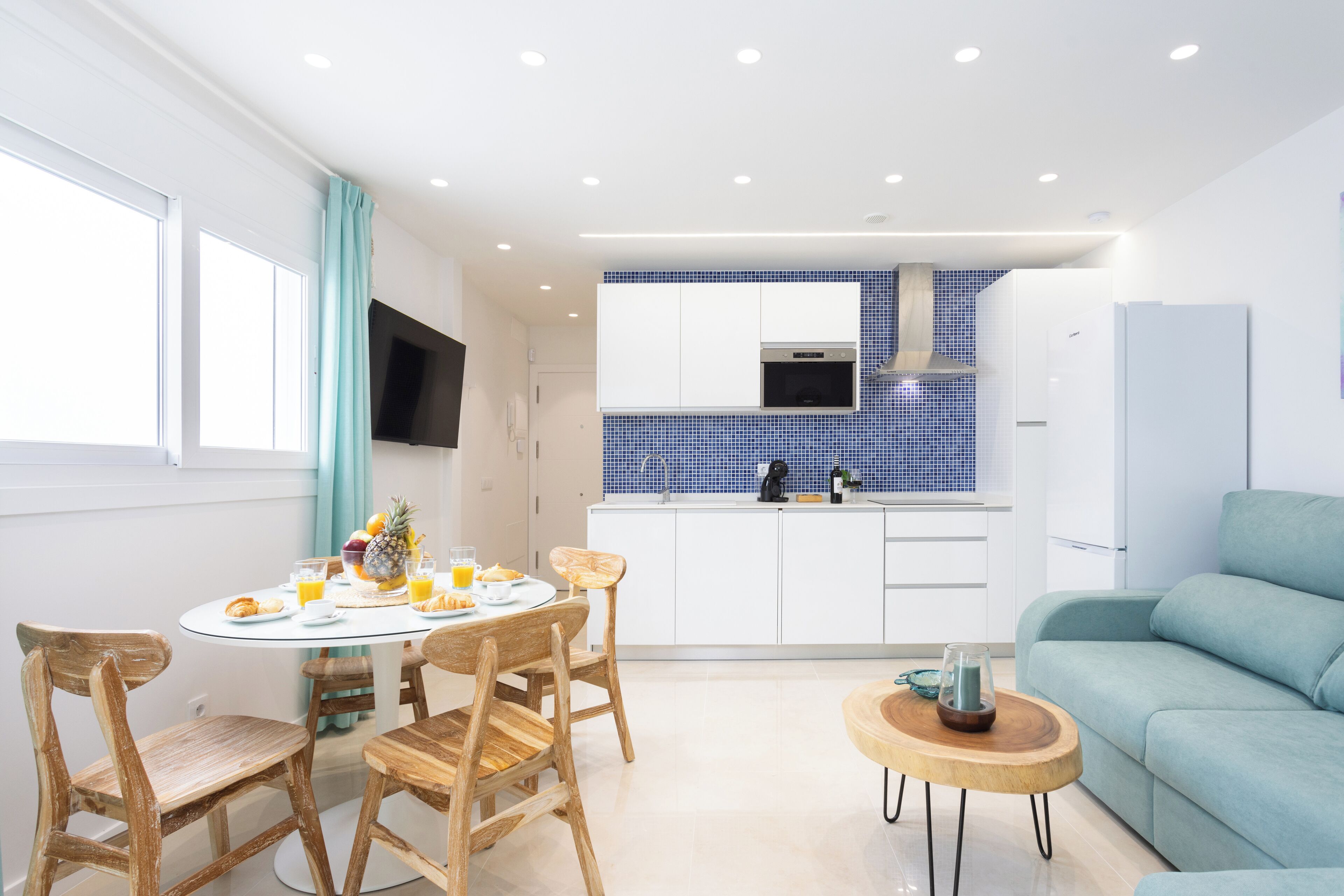 Cocina moderna con armarios blancos, un salpicadero azul y un comedor con sillas de madera y una mesa redonda.