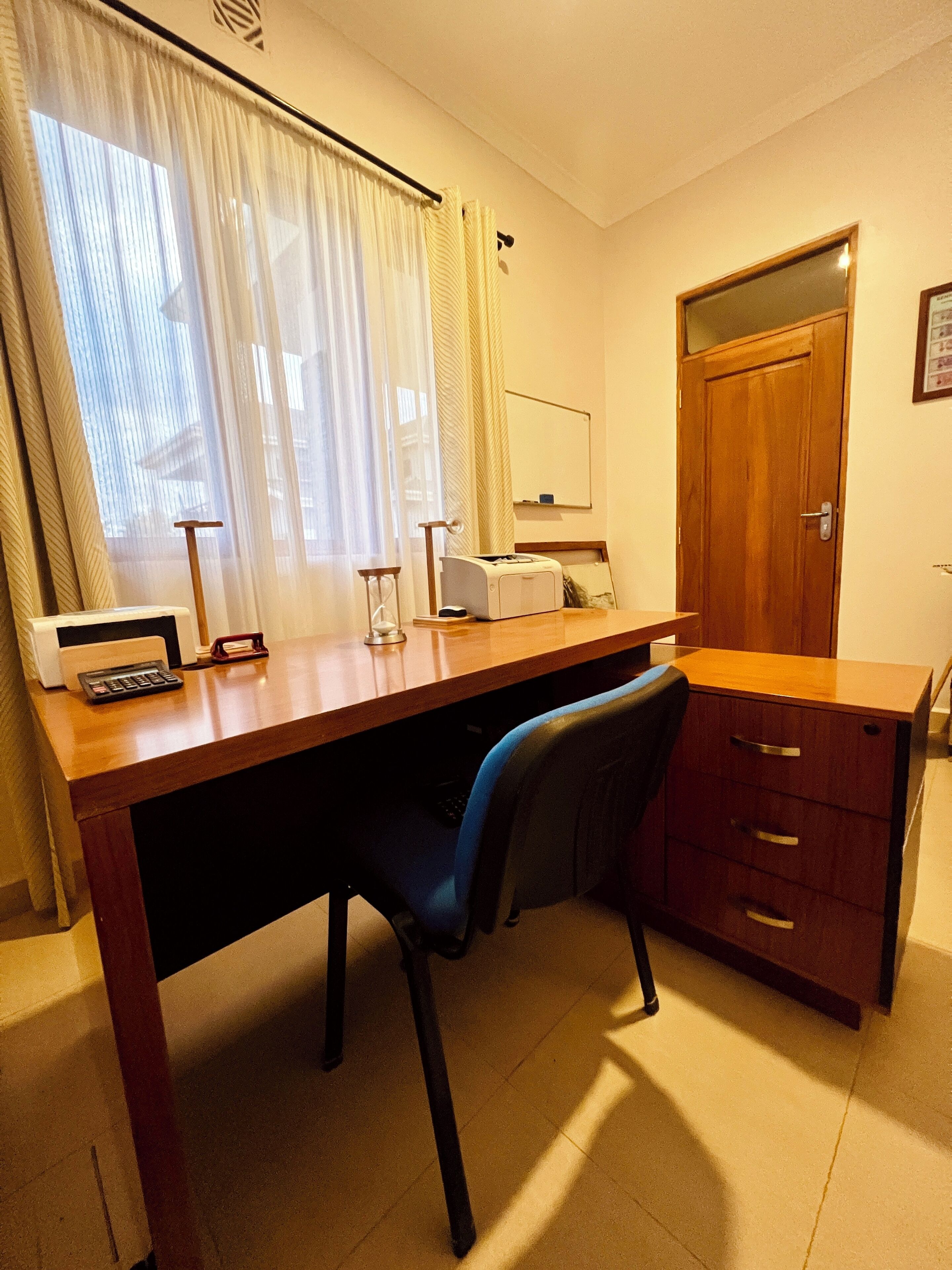 A hotel room with a wooden desk, a chair, a small safe, a mirror, and a window with curtains.