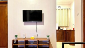 A living room with a flat-screen TV mounted on the wall, a wooden entertainment unit with two potted plants, and a kitchen area visible through an open doorway. - Small White House Njiro (Arusha)