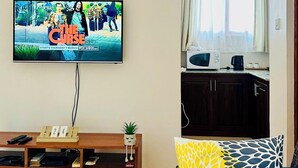 A living room with a TV displaying 'The Curse' poster, a wooden entertainment unit, a sofa with patterned cushions, and a kitchen area with a microwave and kettle. - Small White House Njiro (Arusha)