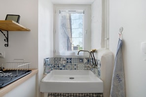 Une petite salle de bain avec un lavabo blanc, une fenêtre avec un rideau, une serviette accrochée au mur et une étagère avec un panier.