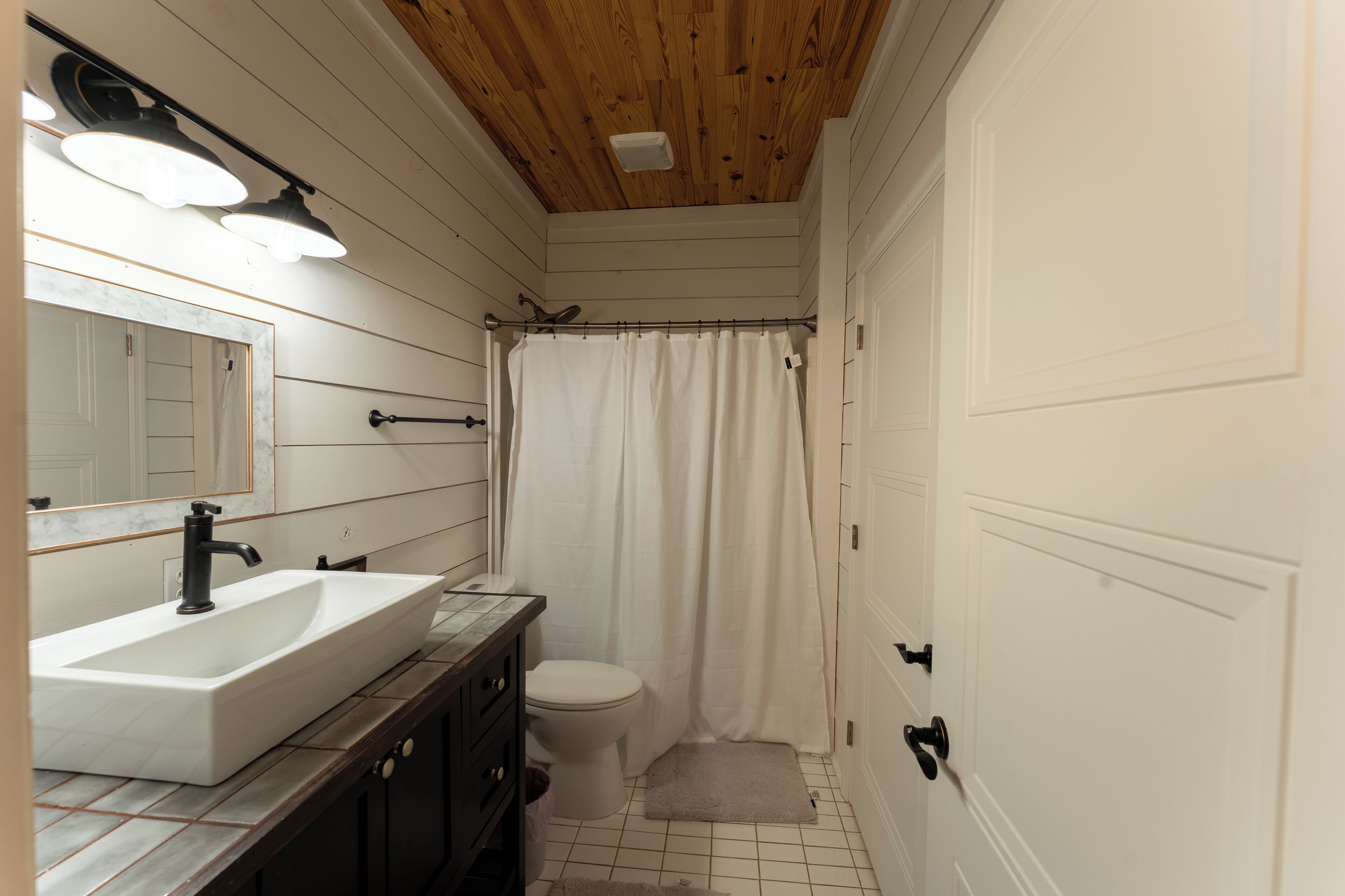 A bathroom with a wooden ceiling, white walls, a white sink, a toilet, and a shower curtain.