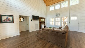 A spacious living room with a wooden ceiling, white paneled walls, a brown leather sofa, a fireplace, and a large window. - Spacious 4-bedroom farmhouse in beautiful Appling with AC (Appling)